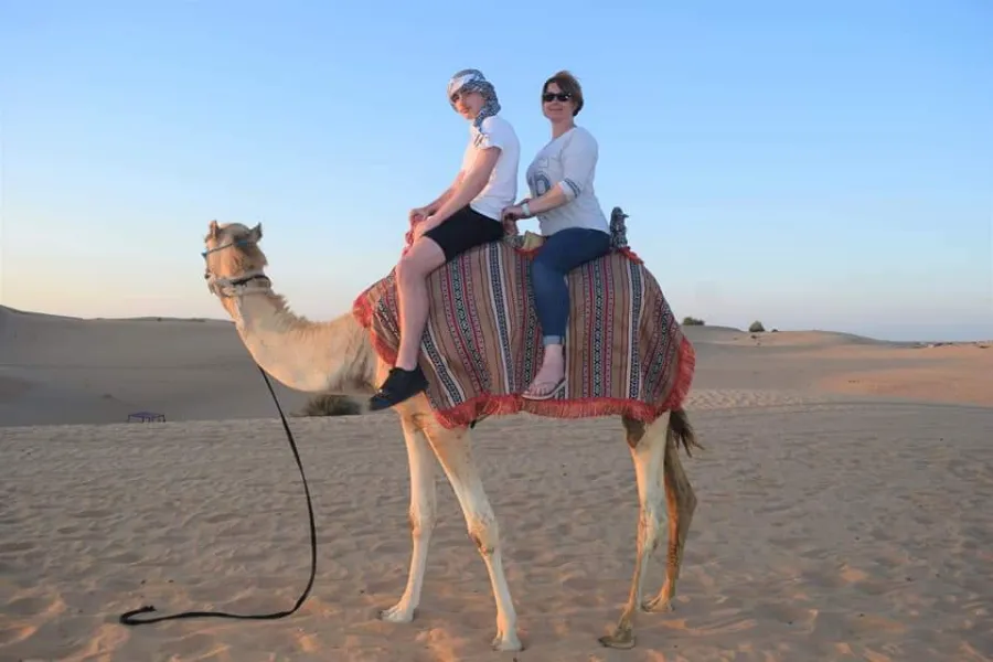 A couple riding a camel together in casual daytime clothing with sandals visible, under a clear blue sky