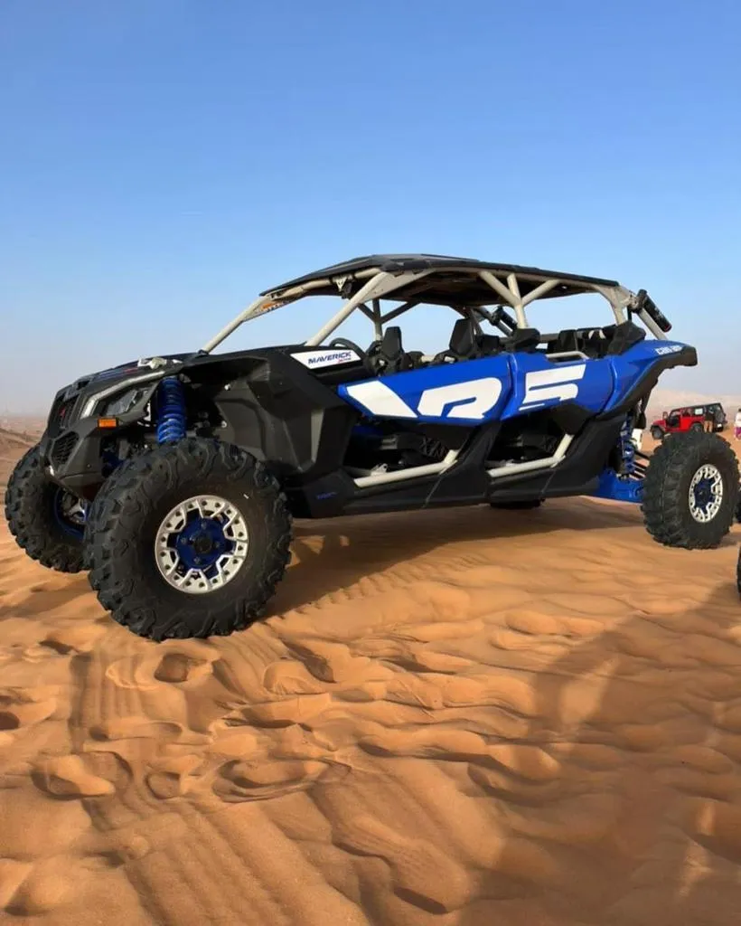 A black and blue off-road quad buggy with a roll cage parked on sand with city buildings visible in the background