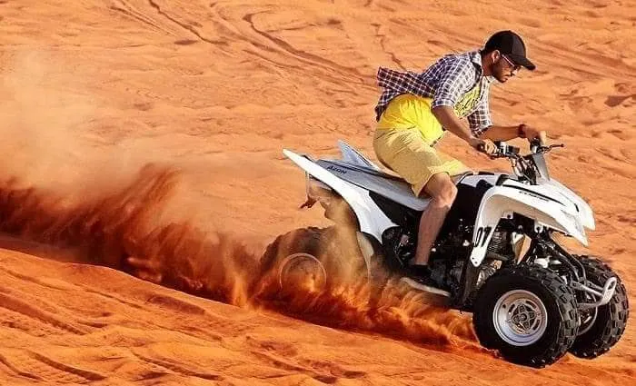 A man in a checked shirt, yellow shorts and a cap riding a quad bike through a spray of orange sand